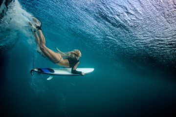 Blonde girl Surfer holding white surf board Diving Duckdive under Big Beautiful Ocean Wave. Turbulent tube with air bubbles and tracks after sea wave crashing. Ripples at water surface with blue sky color.